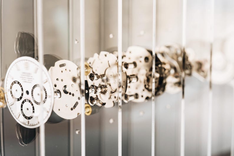 May include: A disassembled watch movement displayed behind vertical glass panels. The white watch face features three black sub-dials and numerical markers. Metallic gears, plates, and internal components are arranged in a linear sequence.