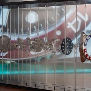 May include: An exploded view of a luxury watch, showcasing its intricate internal components. The watch parts are suspended in a clear display case, revealing the gears, dials, and bezel with red and blue accents. The background features a blurred image of the watch face.