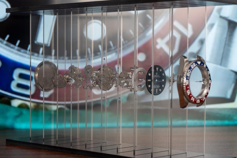 May include: An exploded view of a luxury watch, showcasing its intricate internal components. The watch parts are suspended in a clear display case, revealing the gears, dials, and bezel with red and blue accents. The background features a blurred image of the watch face.