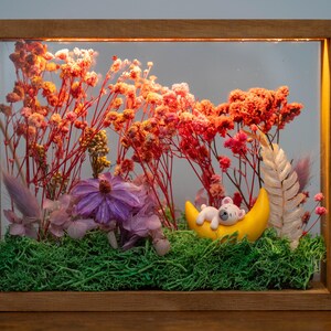 May include: A framed display with dried flowers in red, orange, and pink, with a purple flower and green moss. A yellow crescent moon holds a small, sleeping teddy bear. The wooden frame encloses the scene, creating a miniature diorama.