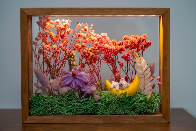 May include: A framed display with dried flowers in red, orange, and pink, with a purple flower and green moss. A yellow crescent moon holds a small, sleeping teddy bear. The wooden frame encloses the scene, creating a miniature diorama.