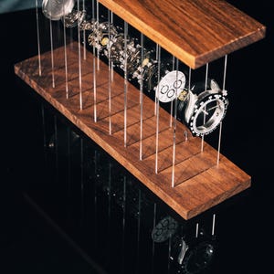 May include: A disassembled wristwatch with its internal mechanical components, including the dial, gears, and casing, suspended between two horizontal dark wood planks by thin, vertical clear acrylic rods, creating an exploded view display.