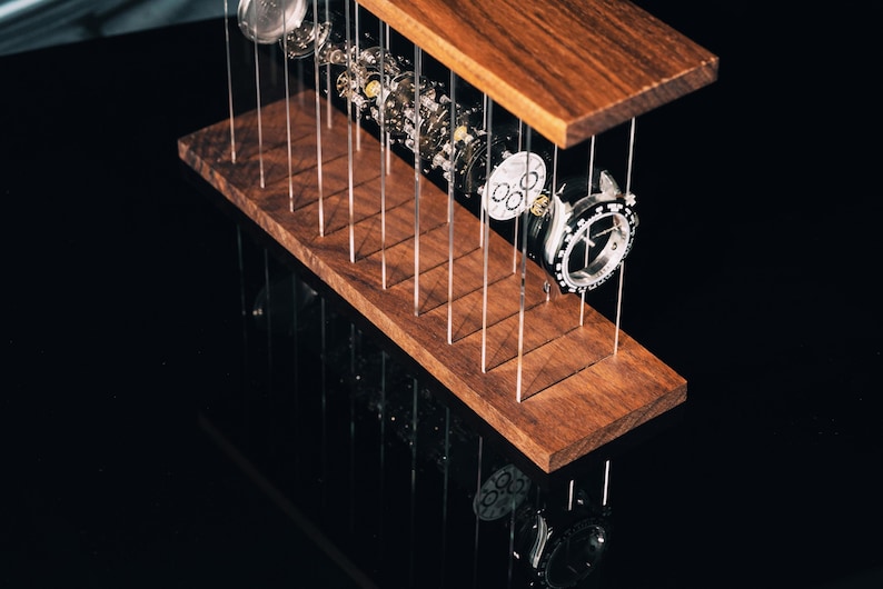 May include: A disassembled wristwatch with its internal mechanical components, including the dial, gears, and casing, suspended between two horizontal dark wood planks by thin, vertical clear acrylic rods, creating an exploded view display.