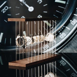 May include: A disassembled watch movement featuring various metallic gears, springs, and components suspended between two horizontal dark wood bars by thin vertical wires, displayed in front of a large black watch face background.