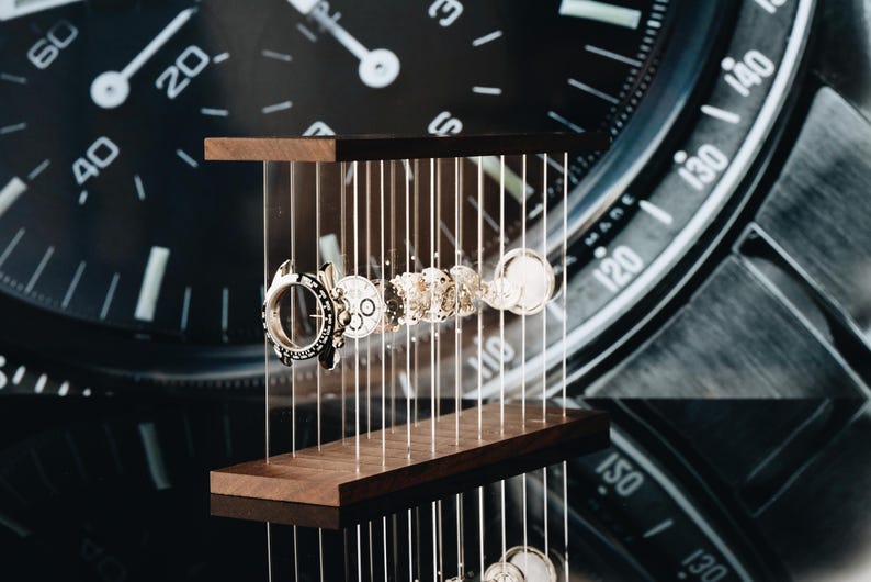 May include: A disassembled watch movement featuring various metallic gears, springs, and components suspended between two horizontal dark wood bars by thin vertical wires, displayed in front of a large black watch face background.