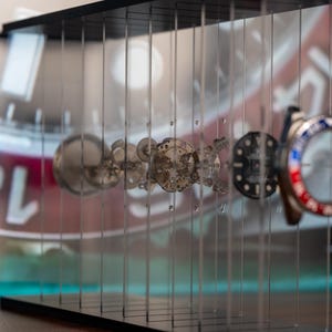 May include: A display of watch components, including gears, dials, and a bezel with red and blue accents. The components are suspended in a clear acrylic case, with a watch face visible in the background.
