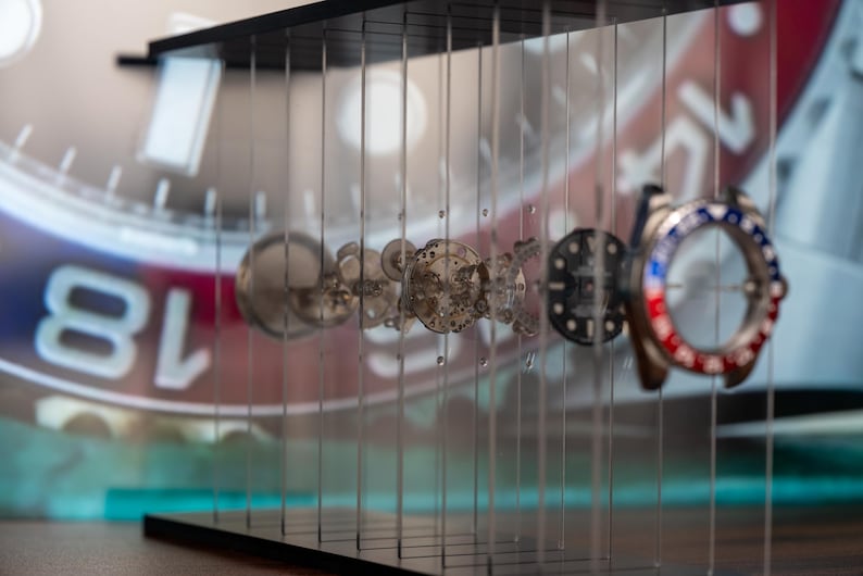 May include: A display of watch components, including gears, dials, and a bezel with red and blue accents. The components are suspended in a clear acrylic case, with a watch face visible in the background.