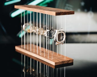 May include: A disassembled wristwatch with its components suspended in a horizontal line between two parallel rectangular wooden planks by thin vertical wires. The watch parts include a silver case, a black dial, and various internal movement gears.