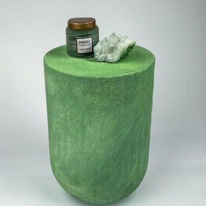 May include: A green candle jar with a gold lid and a white label that reads "Emberly" sits atop a green cylindrical pedestal. A piece of light green crystal is next to the candle. The pedestal is on a white background.