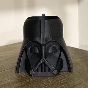 May include: A black plastic Darth Vader head planter. The planter is shaped like Darth Vader's head and has a hole in the top for planting.