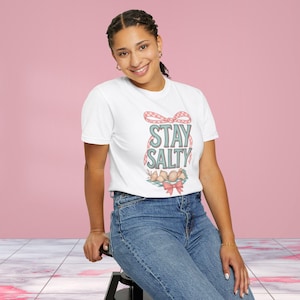 Può includere: T-shirt bianca con la scritta "STAY SALTY" in verde acqua e rosa. Il design include un fiocco e una composizione decorativa di conchiglie e fiocchi. La modella indossa jeans blu.
