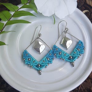 May include: A pair of silver earrings with a blue and turquoise macrame design. The earrings feature a silver geometric pendant.
