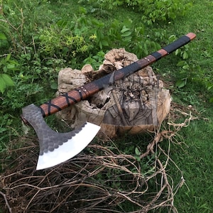 以下が含まれることがあります： 木製の柄と、質感のある銀色の刃を備えた手作りの斧。 柄は黒革で包まれ、革ストラップで固定されています。 斧は、彫刻が施された木の切り株の上に展示されています。