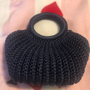 May include: A navy blue crocheted handbag with a circular handle. The bag has a rounded shape with a textured, ribbed design. The handle is a darker shade of navy blue.