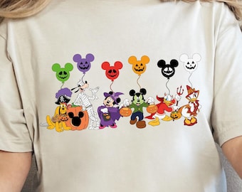 May include: Beige t-shirt featuring a colorful Halloween-themed graphic. The design includes cartoon characters dressed in costumes, holding balloons shaped like Mickey Mouse heads. A pumpkin is also included in the design.