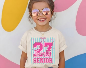 May include: A cream-colored t-shirt featuring a graphic print with the number 27 in large pink block letters, the word Twenty in pink script, and the word SENIOR in pink block letters, accented by a pink bow and light blue vertical stripes.