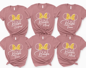 May include: Six dusty rose t-shirts with a Minnie Mouse design. Each shirt features a gold glitter bow and the words "Bride Squad", "Brides Maid", or "Bride Team" in white script, perfect for a bridal party.