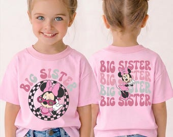 May include: A light pink t-shirt featuring a graphic of Minnie Mouse wearing sunglasses inside a black and white checkered circle on the front, and the text BIG SISTER repeated with a Minnie Mouse illustration on the back.