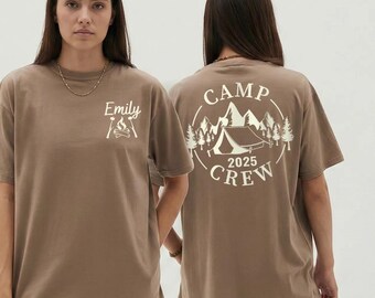 May include: Brown t-shirt with white graphic text and design. The front of the shirt says "Emily" above a campfire graphic. The back of the shirt features a camping scene with the words "CAMP CREW 2025" in a circular design.