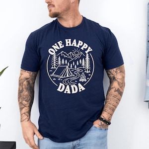 A man wearing a navy blue t-shirt with a graphic design that says "One Happy Dada" and features a camping scene with mountains, pine trees, and a tent.
