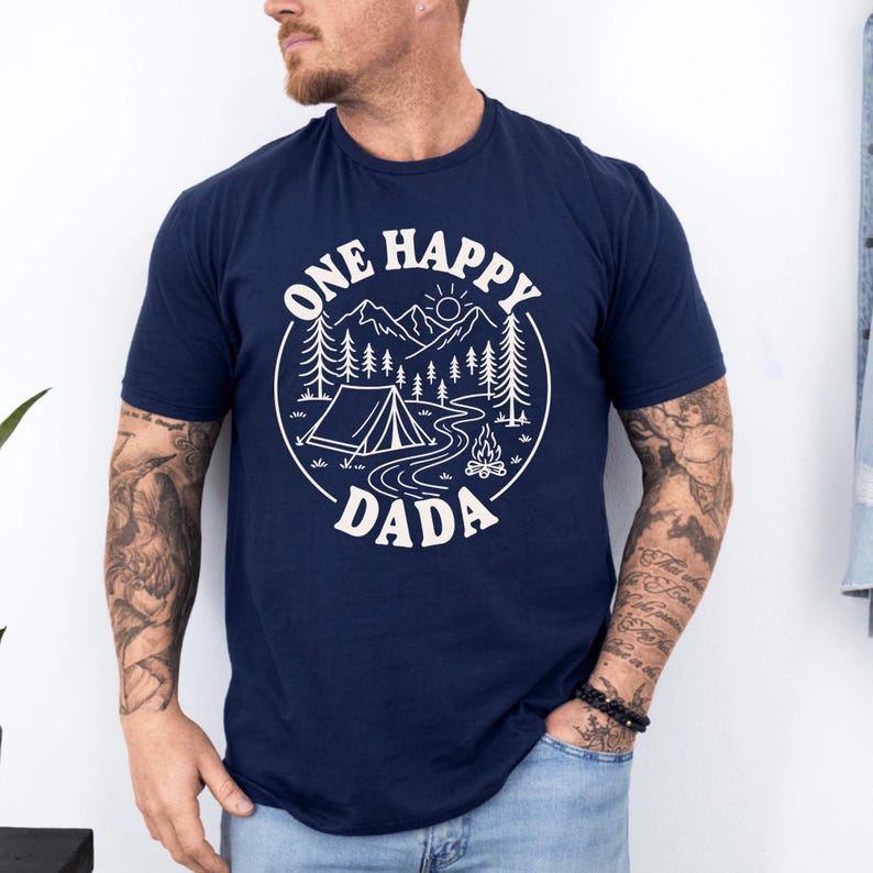 A man wearing a navy blue t-shirt with a graphic design that says "One Happy Dada" and features a camping scene with mountains, pine trees, and a tent.