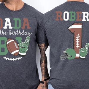 two gray t-shirts with a graphic design on the back. The design includes a football, a number 1, and the text "DADA of the birthday B" and "ROBERT 1".