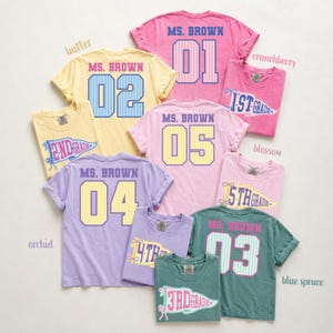 May include: A collection of short-sleeved t-shirts in various pastel colors including pink, yellow, lavender, and blue-green. Each shirt features the text MS. BROWN above a large number or a pennant graphic with grade levels from 1st to 5th grade.