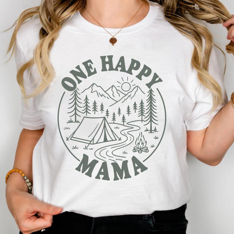 A woman wearing a white t-shirt with a graphic design that says "One Happy Mama" and features a camping scene with a tent, campfire, and mountains.