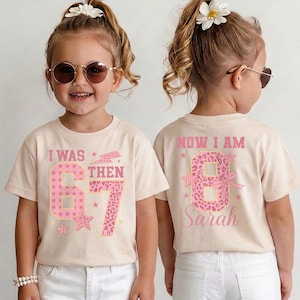 May include: Two children wearing matching beige t-shirts with pink text and graphics. The front of the shirt says "I WAS 67 THEN" with a star and lightning bolt design. The back says "NOW I AM 8 Sarah" with a leopard print number 8.