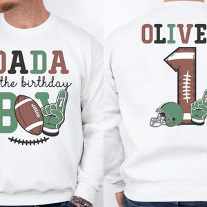 A white sweatshirt with a graphic design featuring a football and the text "DADA of the birthday" and "OLIVER 1" printed on it.