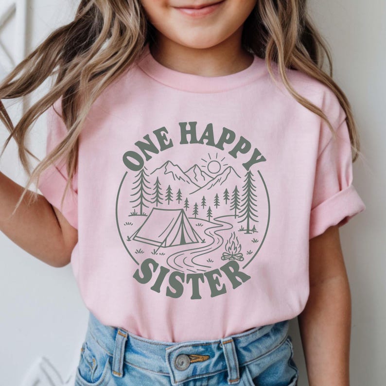A young girl wearing a pink t-shirt with a graphic design of a camping scene and the text "One Happy Sister" printed on it.