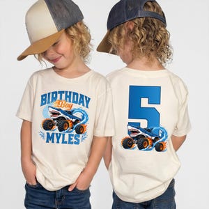 May include: Two cream-colored t-shirts featuring a blue monster truck with shark features. The front shirt displays the text "BIRTHDAY Boy MYLES" in blue and orange. The back shirt displays a large blue number "5" above the same monster truck.