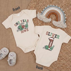 two baby onesies with a football theme, one with the number 1 and the other with the number 1, along with a rainbow, a rainbow-colored afghan, and baby shoes.