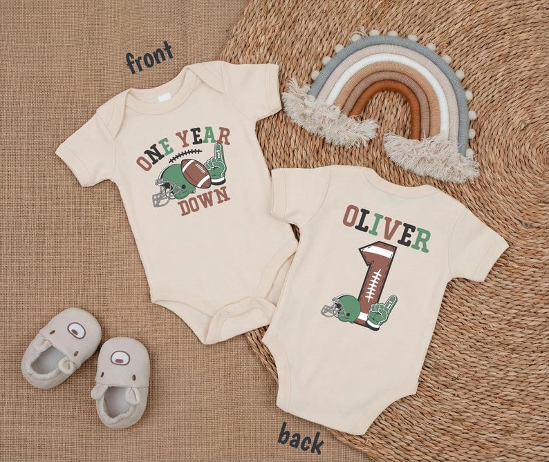 two baby onesies with a football theme, one with the number 1 and the other with the number 1, along with a rainbow, a rainbow-colored afghan, and baby shoes.