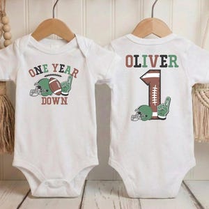 two white baby onesies with a football design and the text "One Year Down" printed on them.