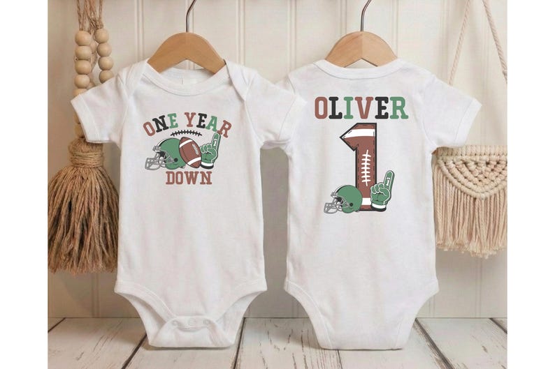 two white baby onesies with a football design and the text "One Year Down" printed on them.