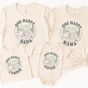 a collection of four beige t-shirts with a graphic design featuring camping and nature elements, along with the text "One Happy Dada" and "One Happy Mama" printed on them.