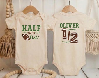 May include: Two beige baby onesies with football-themed graphics. One says "HALF WAY TO one" with a football, the other says "OLIVER 1/2" with a football graphic. Both are on wooden hangers.