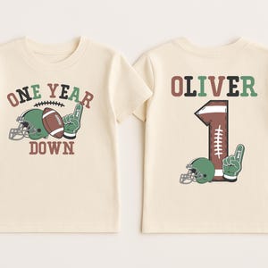 two white t-shirts with a football and a helmet printed on them, along with the text "One Year Down" and the name "Olivier" printed on the back.