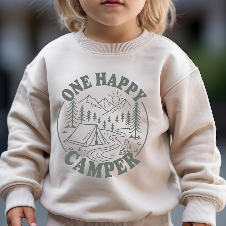 A child wearing a white sweatshirt with a graphic design that says "One Happy Camper" and features a camping scene with a tent, campfire, and mountains.