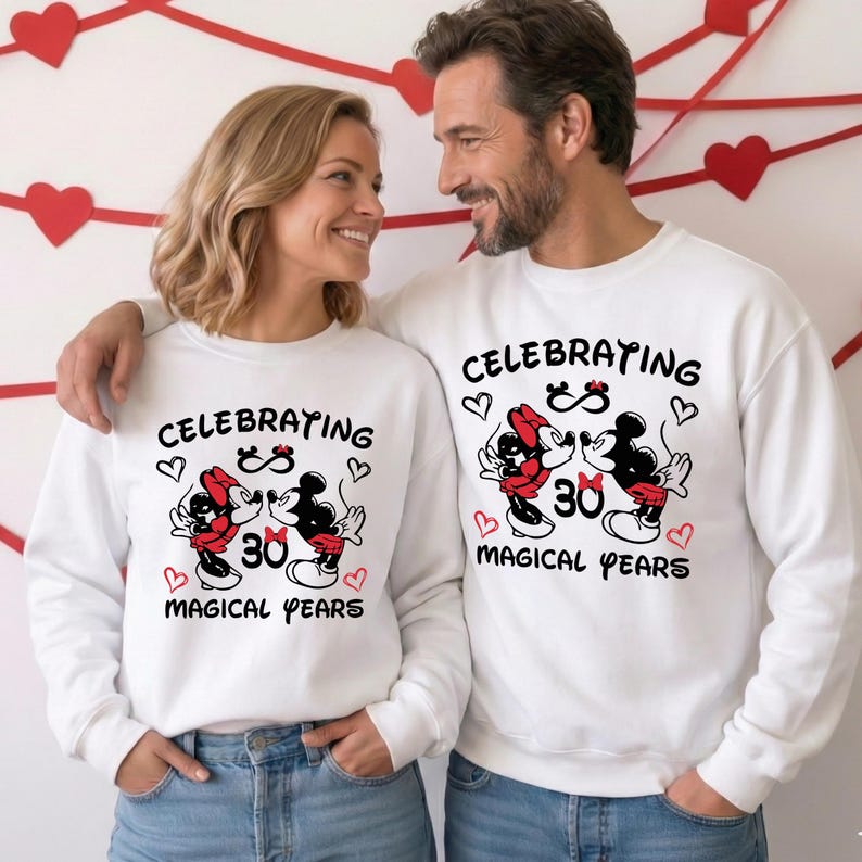 A smiling couple wearing matching white sweatshirts featuring a Disney Disneyland inspired Mickey Minnie 30 Magical Years anniversary design. The graphic shows the iconic cartoon couple kissing with red heart accents and bold milestone lettering. They stand close together in a romantic pose, making this outfit perfect for a Disneyland anniversary trip or magical celebration photoshoot.