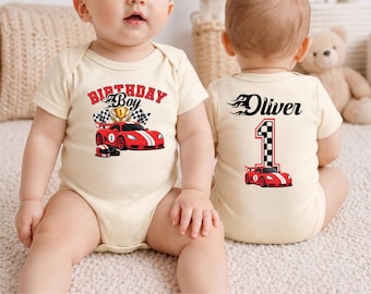 May include: A cream-colored baby bodysuit featuring a red race car graphic, a gold trophy, and the text Birthday Boy on the front. The back displays the name Oliver above a large number one with a checkered flag pattern and a red race car graphic.