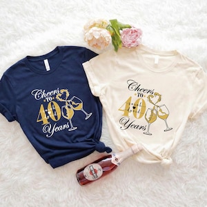 May include: Two t-shirts, one navy blue and one cream, with a gold graphic that says "Cheers to 40 Years." The design includes champagne glasses and a heart. A bottle of red wine is in the foreground.