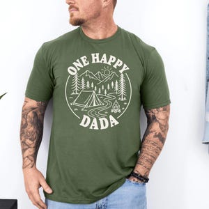 A man wearing a green t-shirt with a graphic design that says "One Happy Dada" and features a camping scene with mountains, pine trees, and a tent.