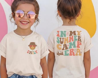 May include: A beige t-shirt featuring a small graphic on the front with a smiley face wearing a bucket hat and the name Emily, and a large graphic on the back with the text In My Summer Camp Era, a camper icon, and various retro-style symbols.