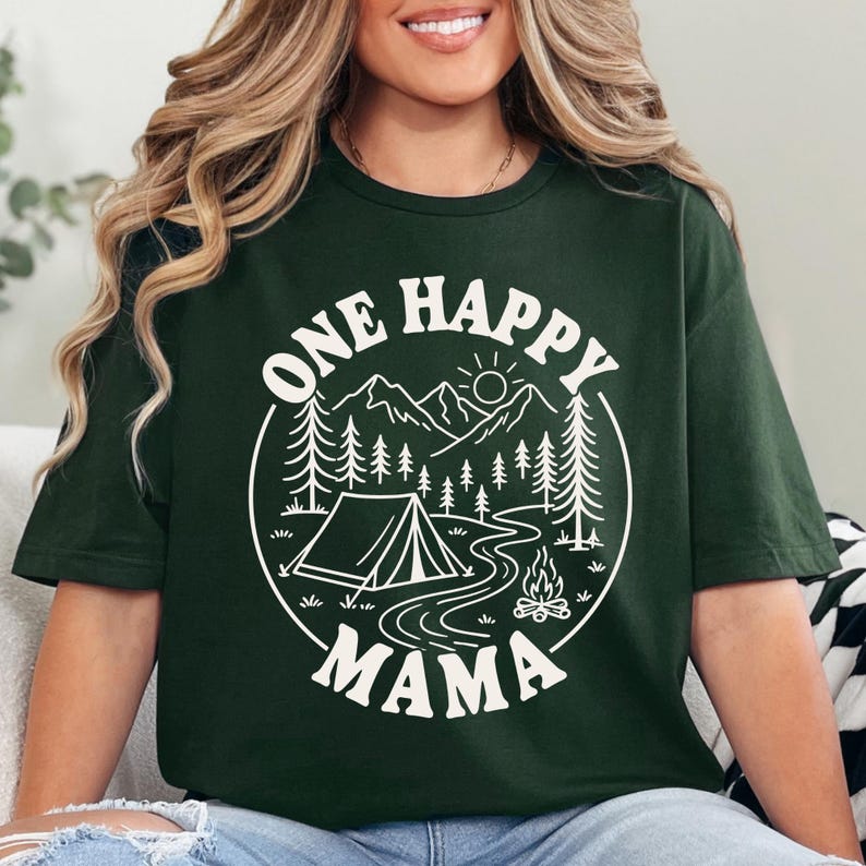 A woman wearing a green t-shirt with a graphic design that says "One Happy Mama" and features a camping scene with mountains, pine trees, and a campfire.