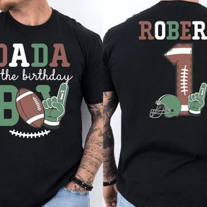 A black t-shirt with a graphic design featuring a football and the text "DADA of the birthday B1" printed on it.