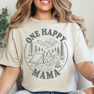 A woman wearing a t-shirt with a graphic design of a camping scene and the text "One Happy Mama" printed on it.