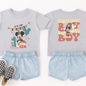 a pair of children's clothing items, including a gray t-shirt with a Mickey Mouse design and a pair of denim shorts, along with a pair of black and white checkered shoes.