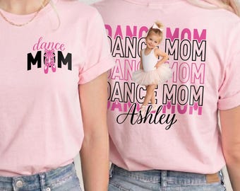 May include: A light pink t-shirt featuring a front graphic with the text dance MOM and pink ballet slippers. The back displays the text DANCE MOM in repeating rows, a photograph of a young child in a white tutu, and the name Ashley in script.